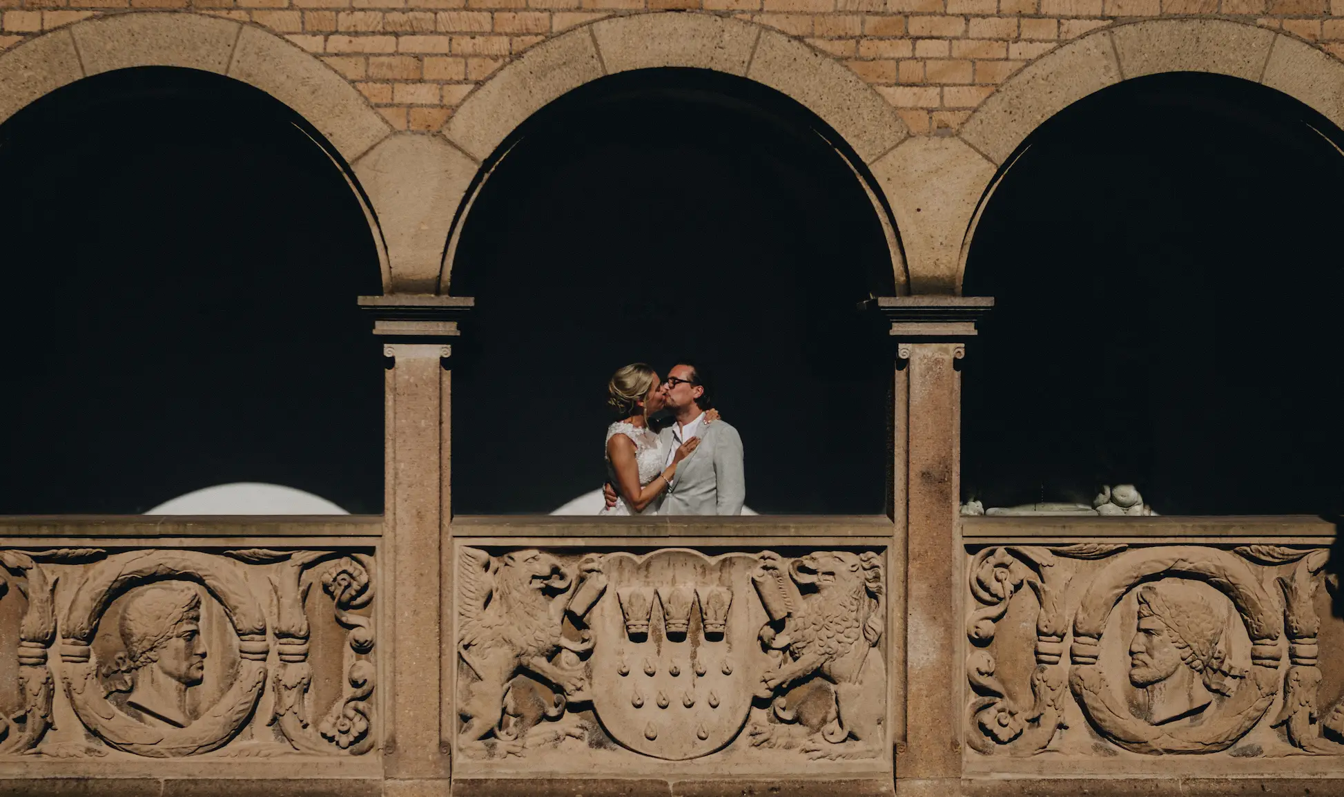 Brautpaar küsst sich liebevoll unter einer historischen Arkade in Köln, vom Sonnenlicht sanft beleuchtet – unvergesslicher Moment der Hochzeit.