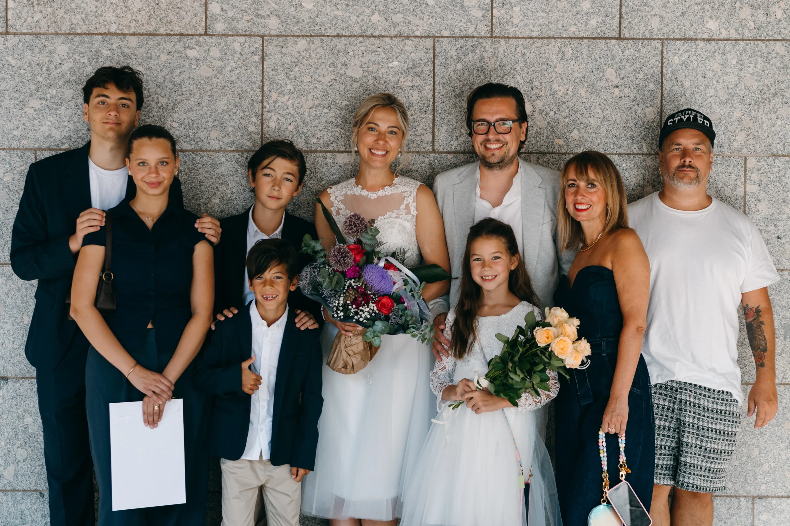 Ein künstlerisches Hochzeitsfoto vor einer Steinmauer in Köln: Das Brautpaar lächelt glücklich inmitten ihrer Familie und Hochzeitsgäste. Die Braut trägt ein elegantes weißes Kleid und hält einen bunten Blumenstrauß, während die Gäste stilvoll in Anzügen und Abendkleidung gekleidet sind.