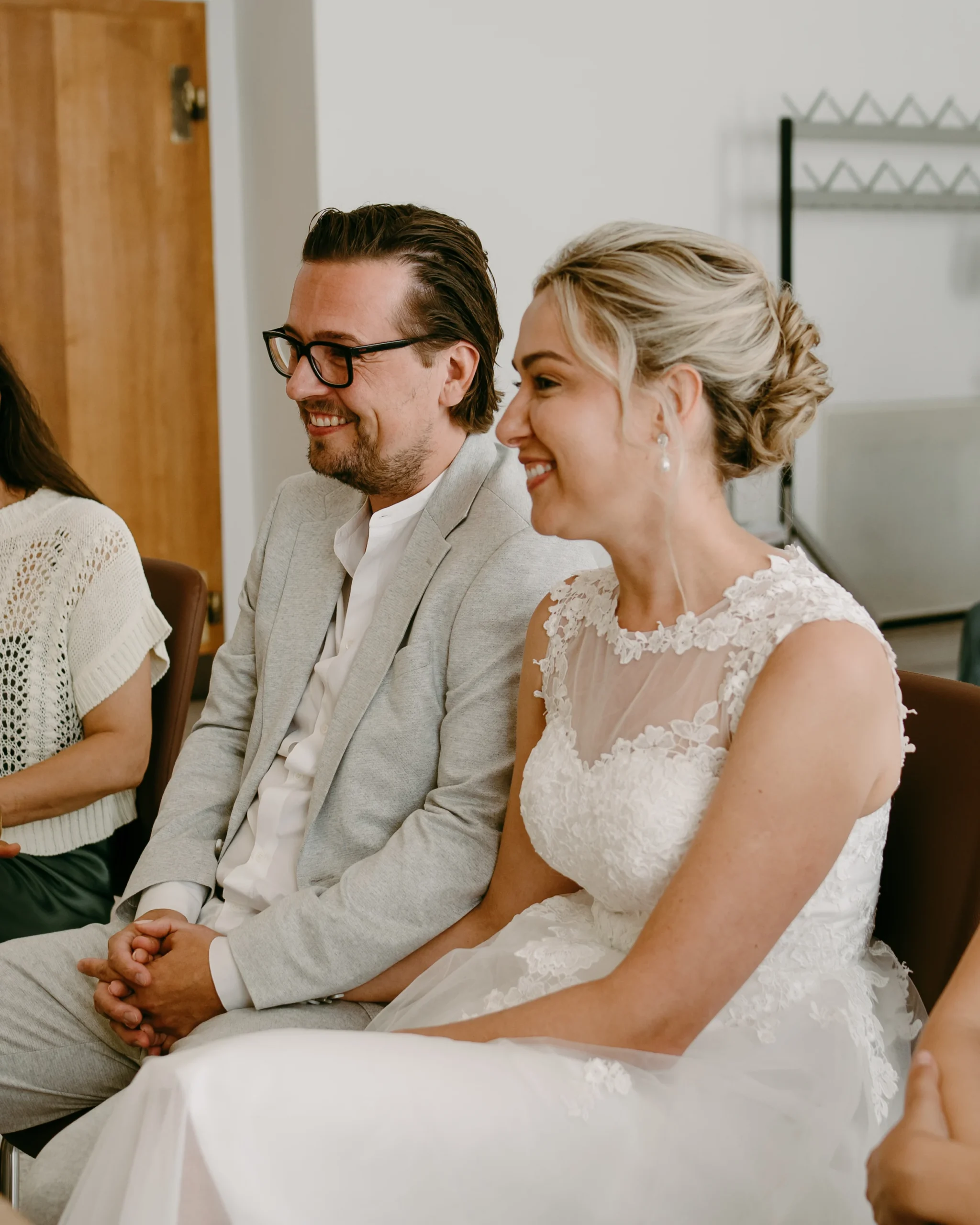 Nahaufnahme eines glücklichen, lächelnden Brautpaares während ihrer standesamtlichen Trauung im Kölner Standesamt, mit Fokus auf die Braut in einem Spitzenkleid und den Bräutigam mit Brille.