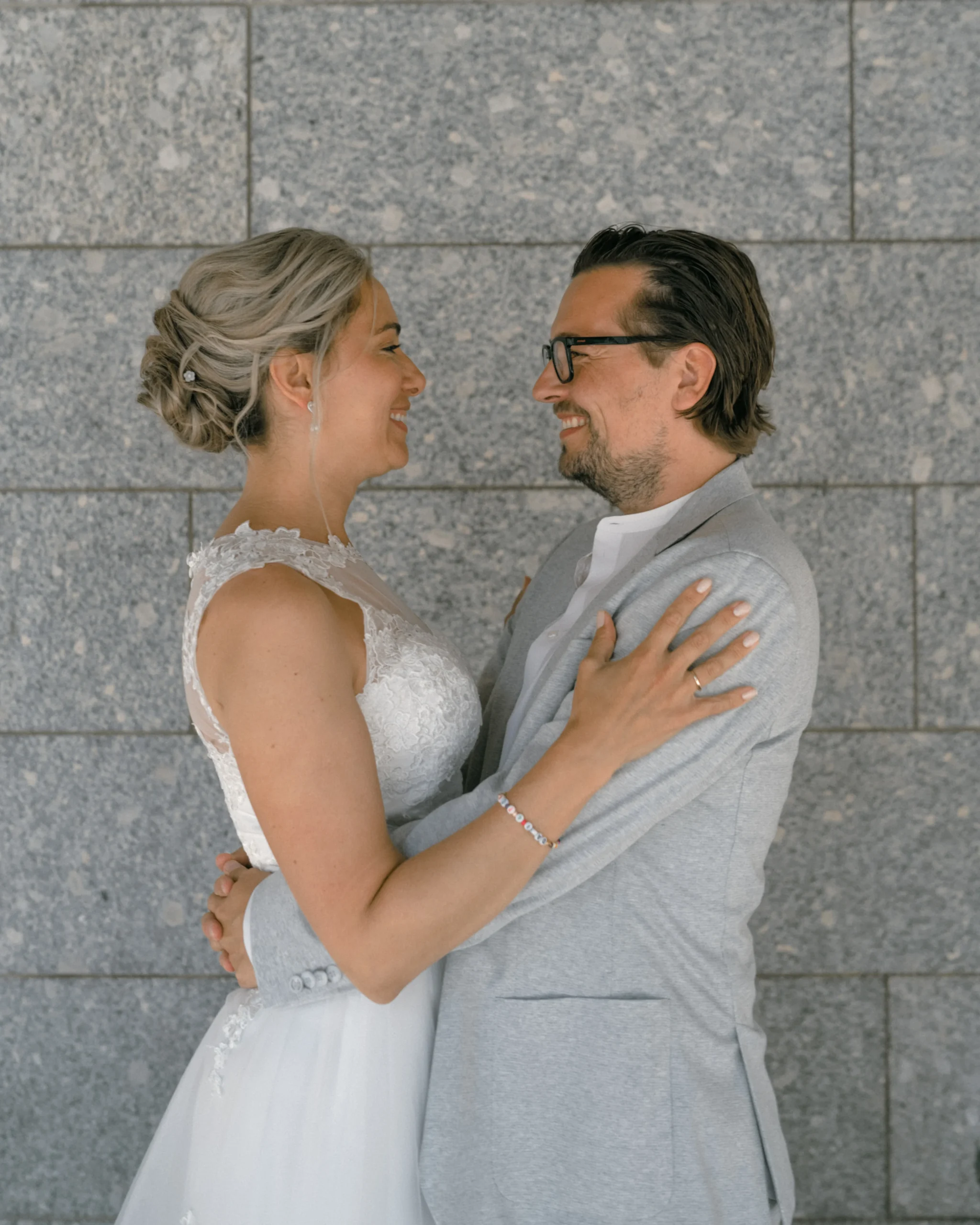 Eine authentische Hochzeitsaufnahme in Köln: Braut und Bräutigam schauen sich tief in die Augen und lächeln, während sie sich vor einer modernen grauen Steinwand umarmen. Die Braut trägt eine elegante Hochsteckfrisur und ein Spitzenkleid, der Bräutigam einen hellgrauen Anzug.
