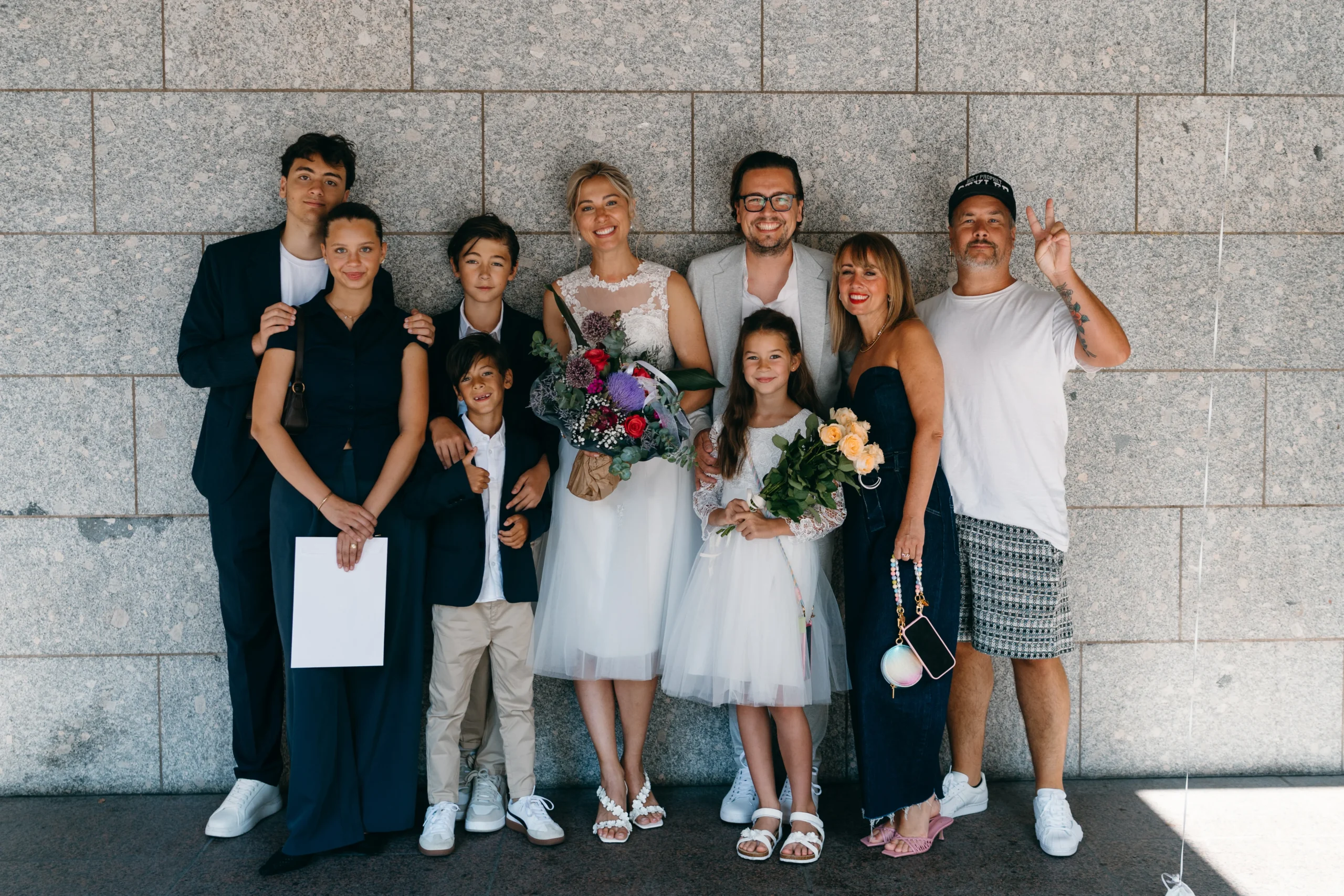 Authentisches Hochzeitsgruppenfoto mit Brautpaar und Gästen in entspannter Atmosphäre vor einer modernen Wand in Köln.
