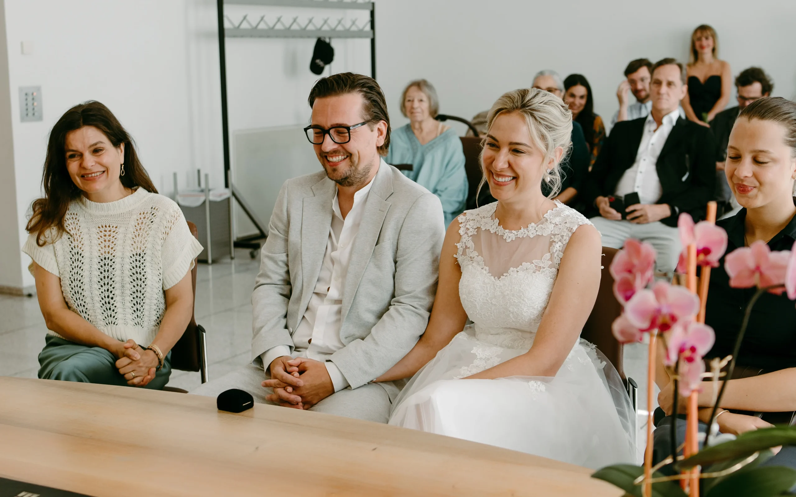 Glückliches Brautpaar sitzt während der standesamtlichen Trauung im Kölner Standesamt mit Trauzeugen am Standesamtstisch und Gästen im Hintergrund, in einem hellen, modernen Trauzimmer.