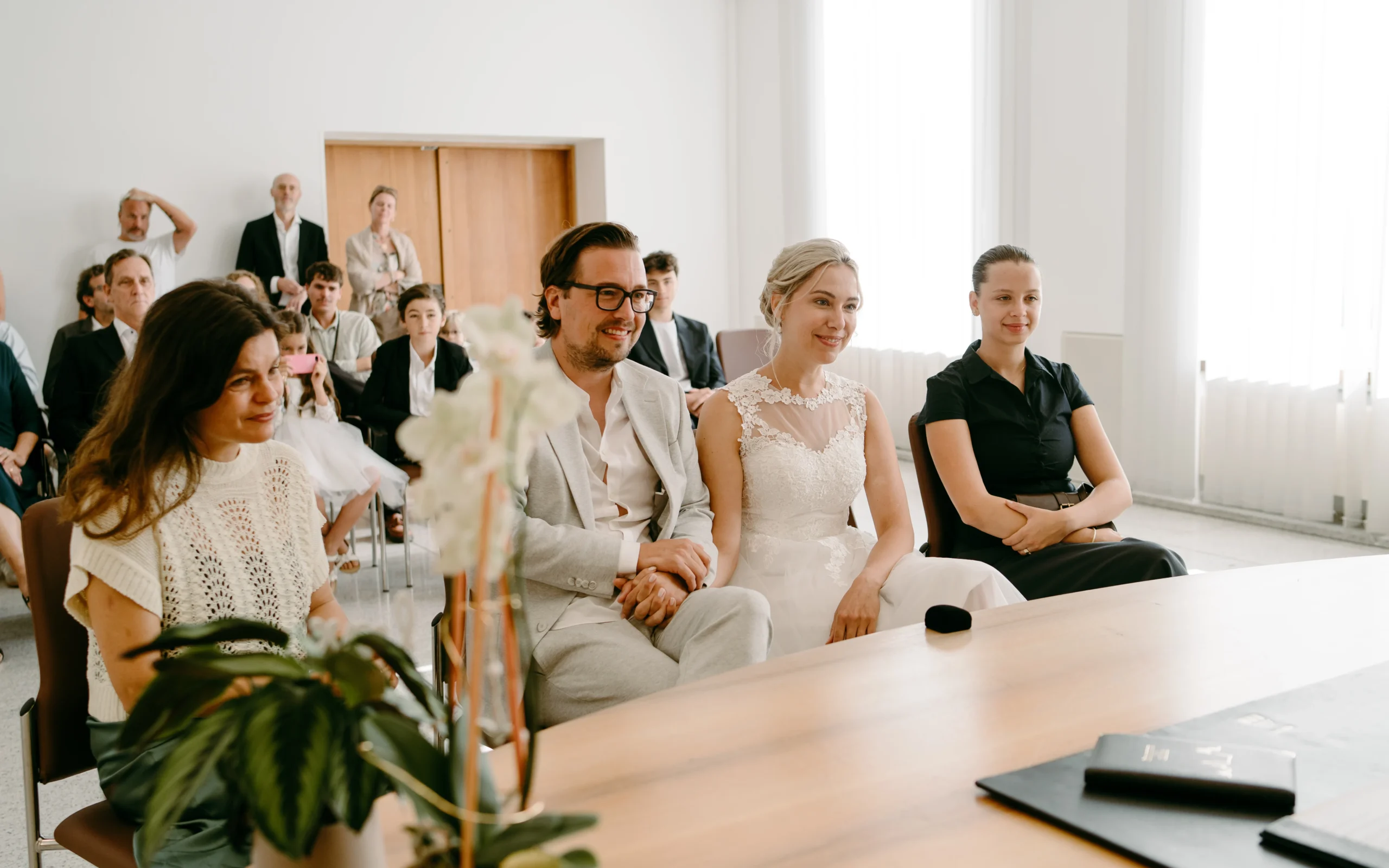 Seitenansicht der Hochzeitszeremonie im Standesamt Köln: Das Brautpaar sitzt mit seinen Trauzeugen am Standesamtstisch in einem hellen, modernen Trauzimmer.