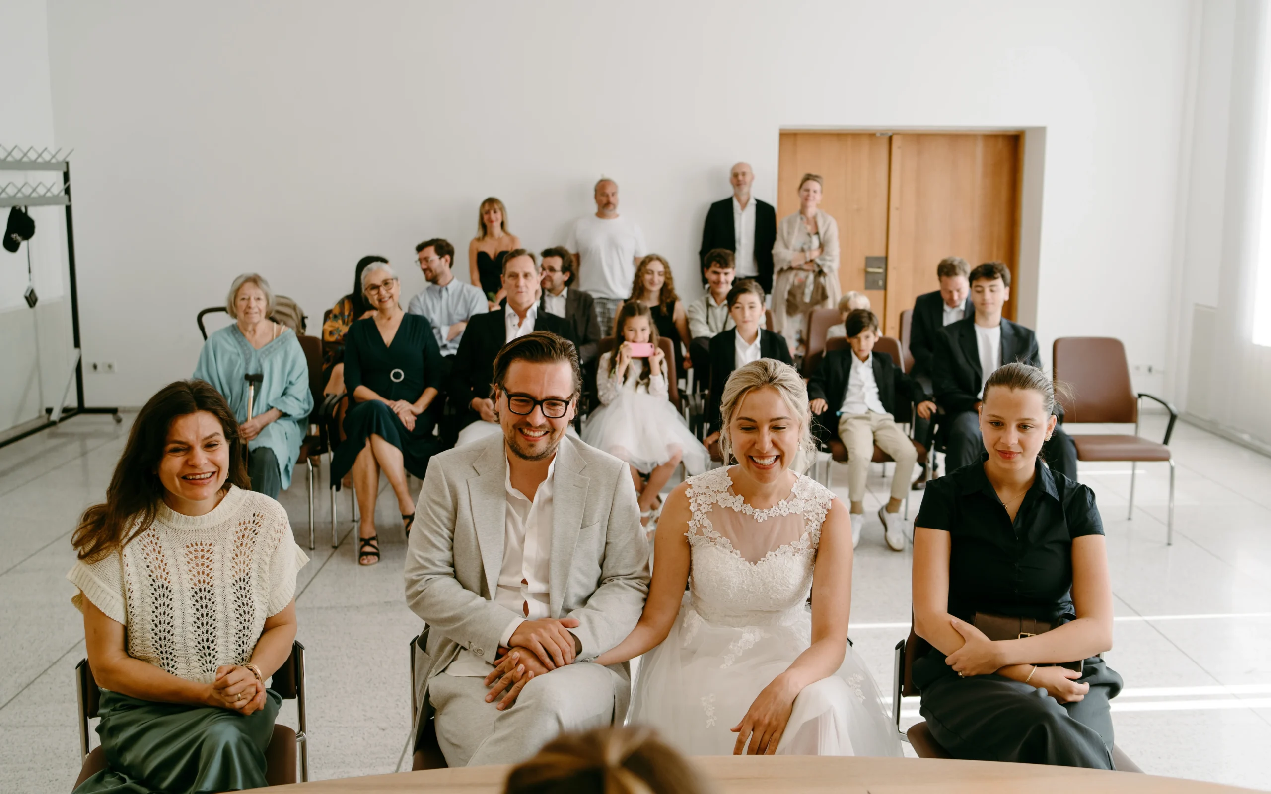 Glückliches Brautpaar sitzt während der standesamtlichen Trauung im Kölner Standesamt zusammen mit Hochzeitsgästen in einem hellen, modernen Saal.