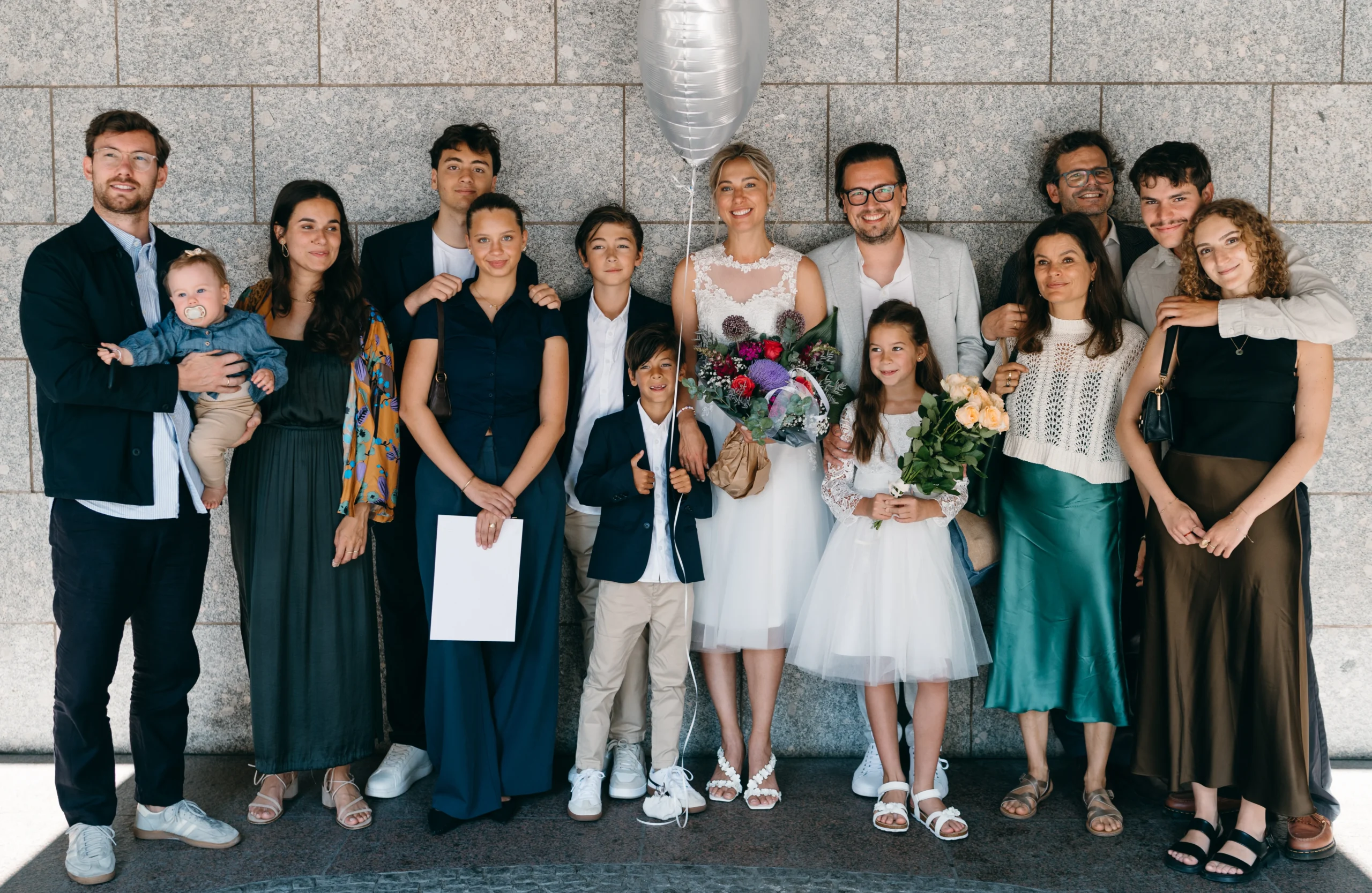 Großes Hochzeitsgruppenfoto mit Brautpaar, Freunden und Familie vor einer Steinwand in Köln.