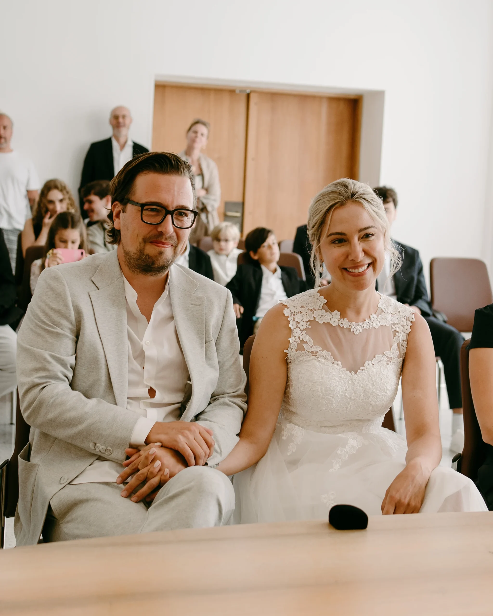 Nahaufnahme des glücklichen Brautpaares, Händchen haltend während ihrer standesamtlichen Trauung im Kölner Standesamt, mit Hochzeitsgästen und modernem Dekor im Hintergrund.
