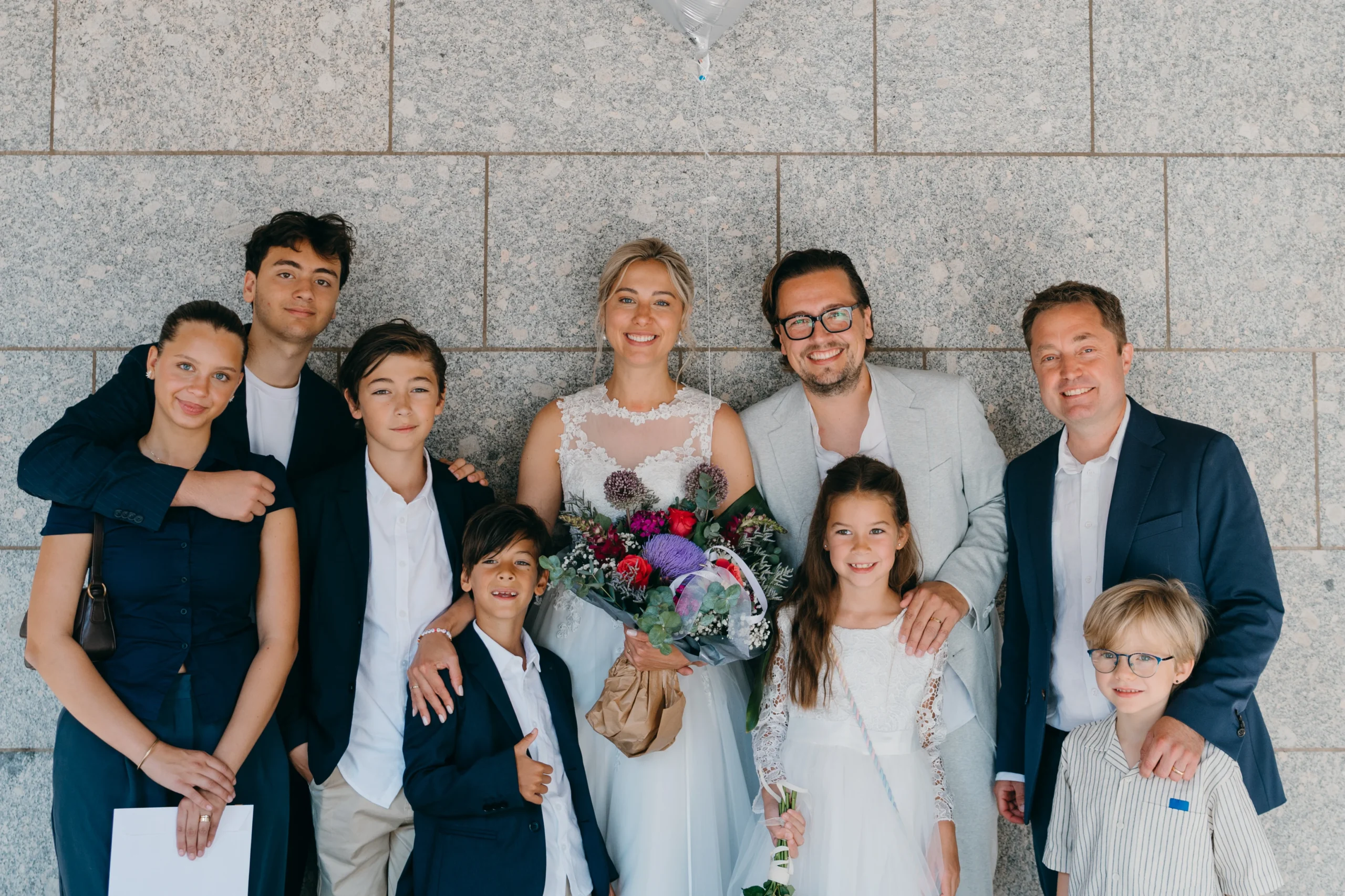 Fröhliche Hochzeitsgesellschaft mit Brautpaar und Kindern vor einer modernen Steinfassade in Köln.