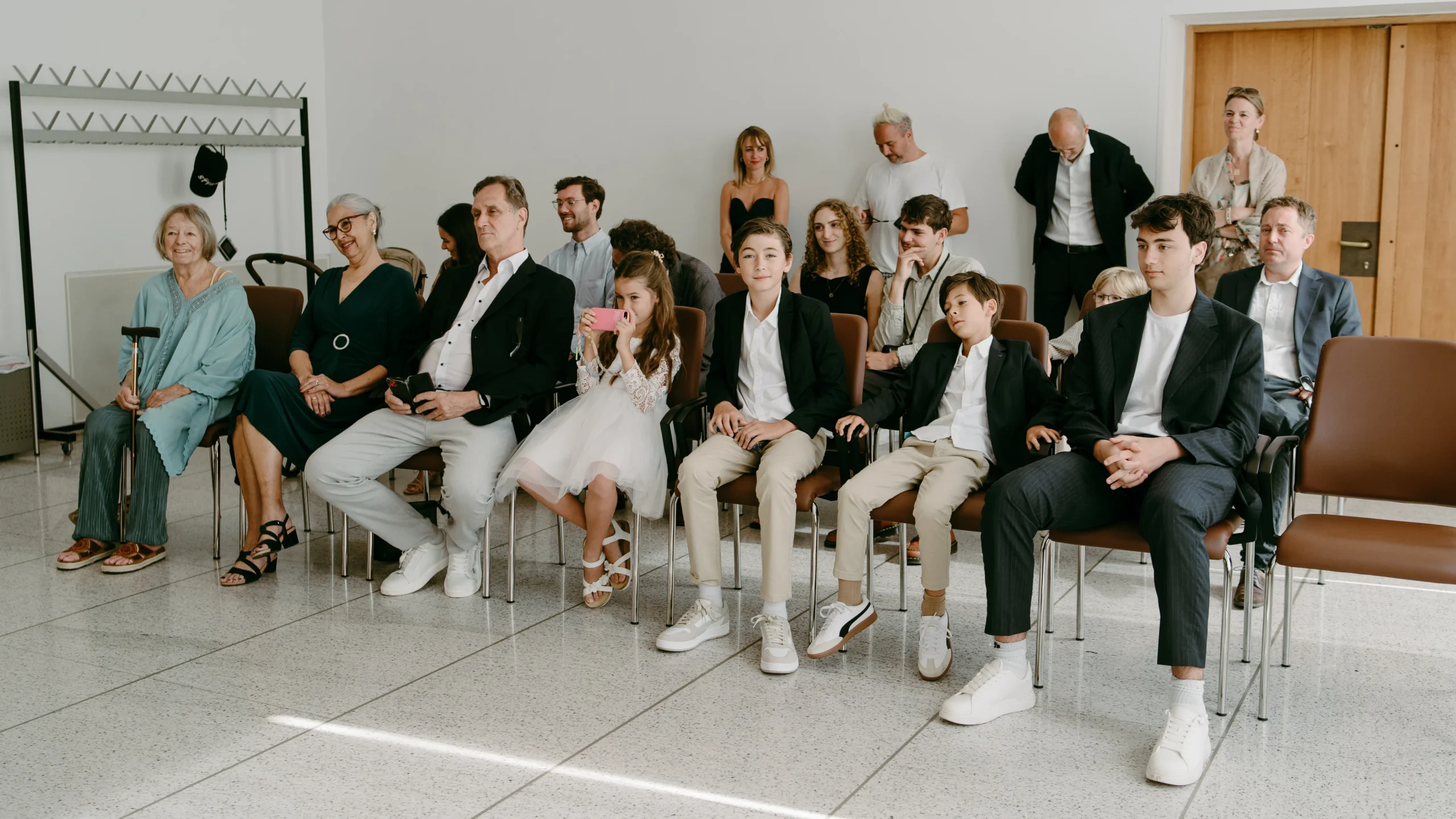 Hochzeitsgäste sitzen während einer intimen standesamtlichen Trauung im Kölner Standesamt auf modernen braunen Stühlen in einem hellen, minimalistischen Trauzimmer.