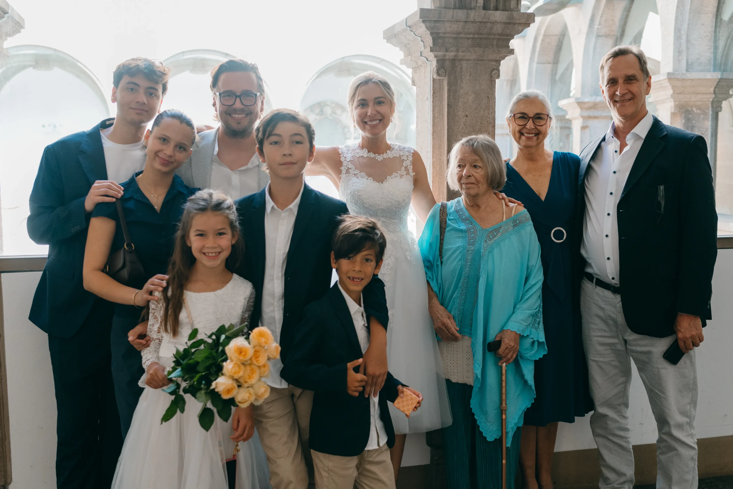 Ein Gruppenfoto eines frisch verheirateten Paares und seiner Familien, die nach ihrer Hochzeitszeremonie auf einem historischen Kreuzgang in Köln stehen. Die Braut trägt ein weißes Kleid und der Bräutigam einen Anzug. Die Gäste sind festlich gekleidet und lächeln.